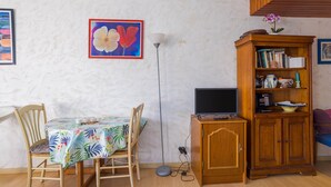 Une pièce avec un meuble en bois, une table recouverte d’une nappe à motifs floraux, un lampadaire et un tableau accroché au mur.