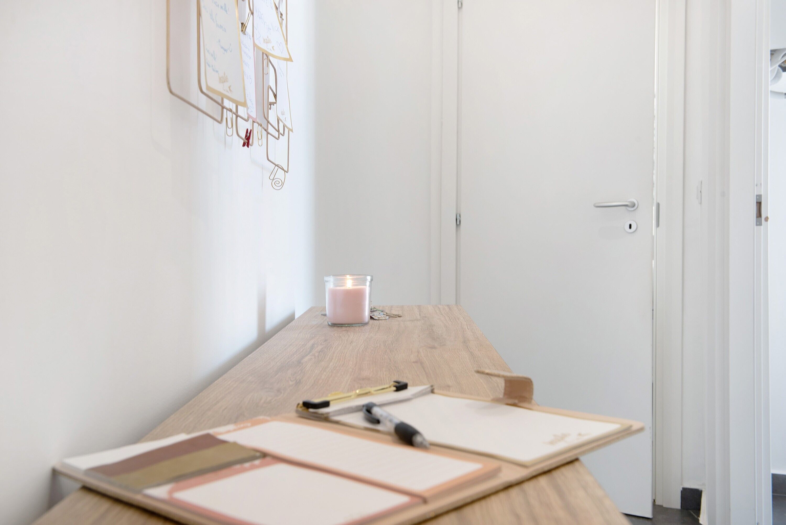 Una scrivania in legno con una candela, una penna e documenti in una stanza con pareti bianche e una porta.