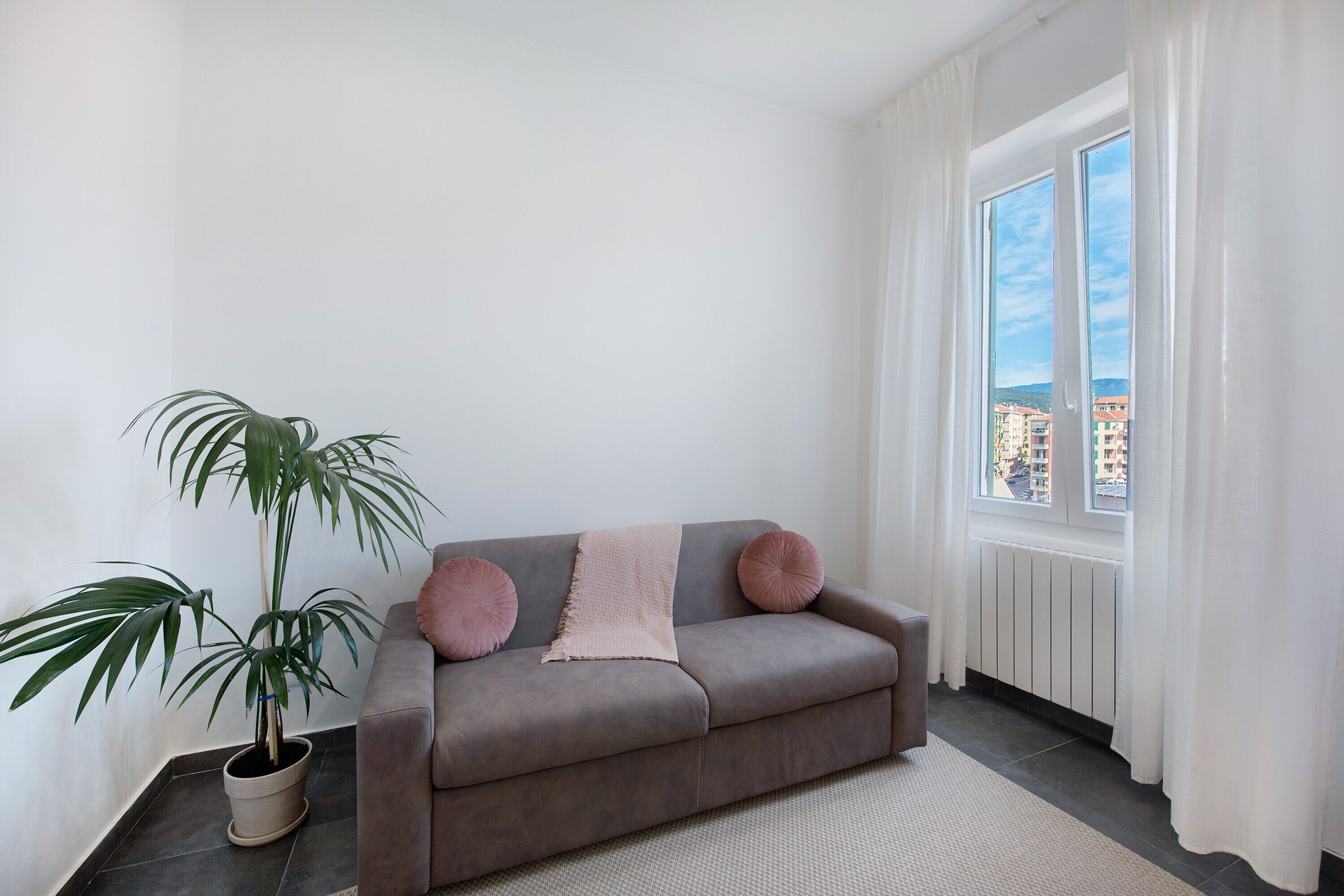 Een grijze bank met een beige deken en twee roze kussens, een potplant en een uitzicht op gebouwen door een raam met witte gordijnen.