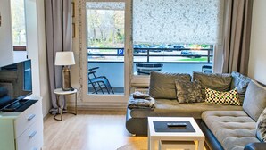 A living room with a sofa, a coffee table, a TV, a lamp, and a view of a parking area through a window. - Koje 44 by Interhome (Damp)