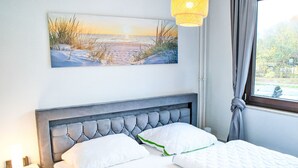 A bedroom with a bed, a nightstand, a lamp, and a painting of a beach scene. - Koje 44 by Interhome (Damp)