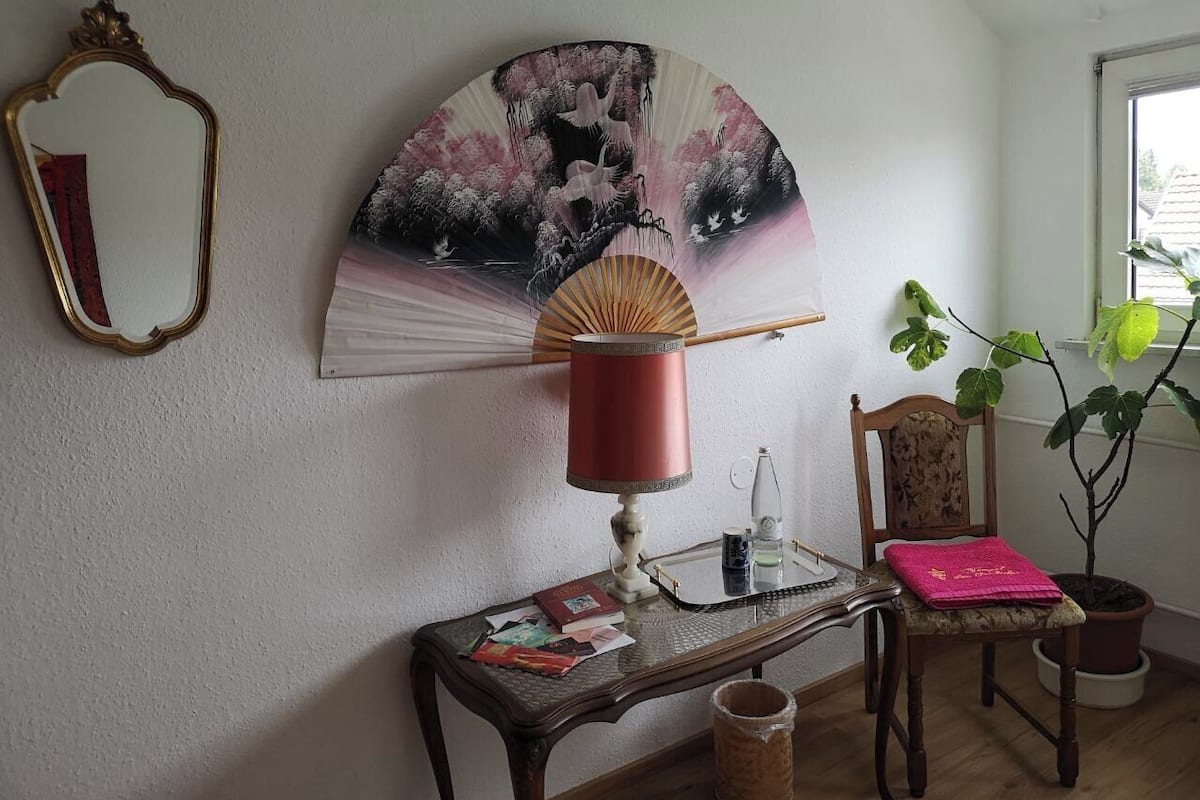 A room with a wooden table, a red lamp, a fan, a mirror, a chair, and a potted plant.