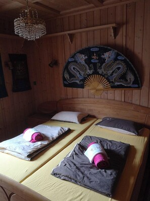 A wooden cabin room with two beds, each with a pillow and a rolled towel. A decorative fan and a chandelier are visible on the wall. - Seminarhaus Arya Maitreya (Kaufbeuren)