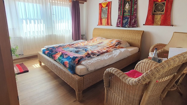 A room with a bed, wicker furniture, and decorative wall hangings. - Seminarhaus Arya Maitreya (Kaufbeuren)