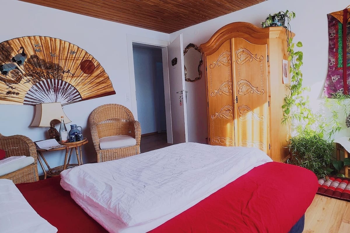 A hotel room with a bed, two wicker chairs, a wooden wardrobe, a wall fan, and a decorative painting.