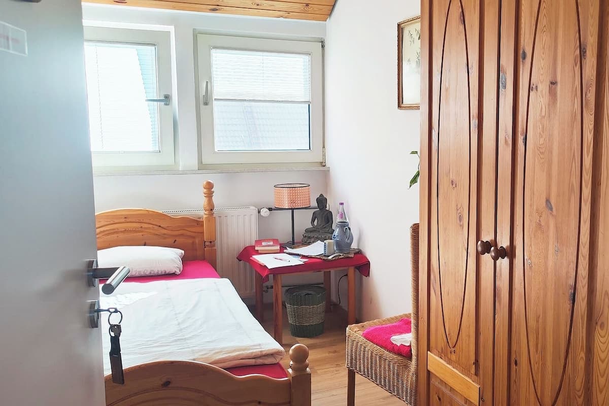 A small bedroom with a wooden wardrobe, a bed with a wooden headboard, a desk with a chair, and a window with blinds.
