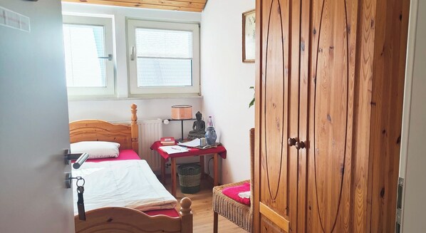 A small bedroom with a wooden wardrobe, a bed with a wooden headboard, a desk with a chair, and a window with blinds. - Seminarhaus Arya Maitreya (Kaufbeuren)