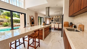 A modern kitchen with a large island, bar stools, a built-in oven, and a view of a swimming pool. - Villa Alexa: Experience the Adventure! (Akumal)