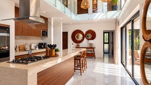 A modern kitchen with a central island, stainless steel appliances, and a glass railing overlooking a dining area. - Villa Alexa: Experience the Adventure! (Akumal)