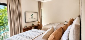A bedroom with a bed, pillows, a bedside table, a framed picture, and a view of greenery outside.