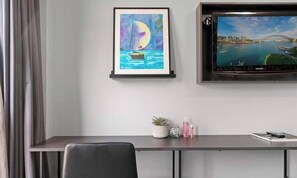 Huoneessa on työpöytä, kehystetty maalaus, jossa on kuvattuna purjevene, televisio, jolta näkyy kaupunkimaisema, ja pieni kasvi työpöydällä.