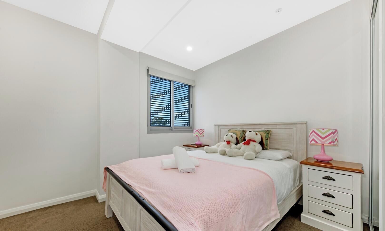 A bedroom with a bed, two bedside tables, a window with blinds, and a nightstand with a lamp.