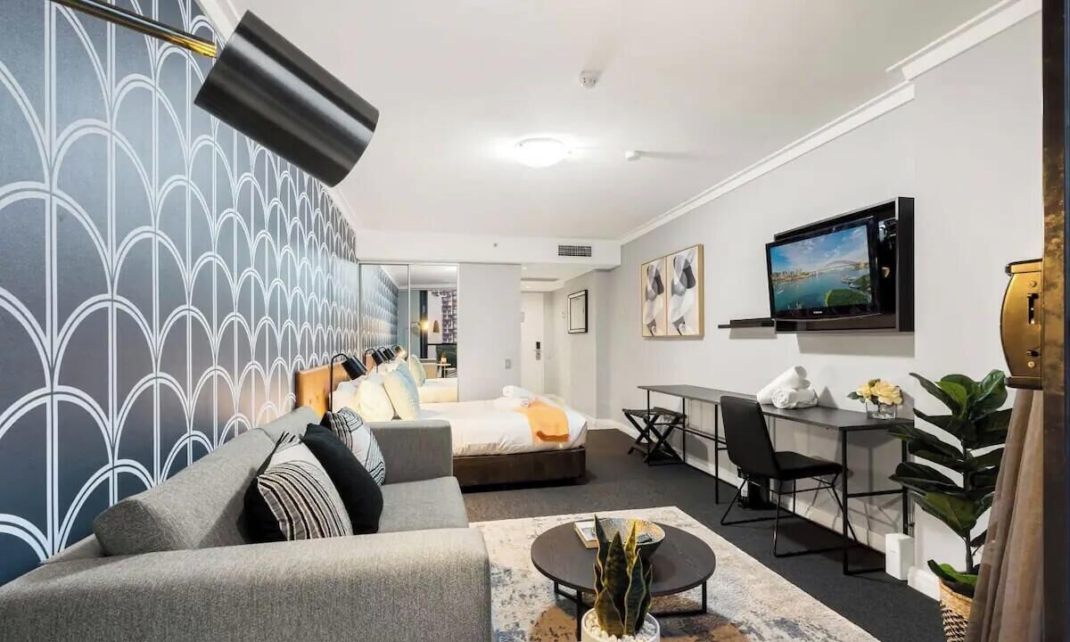 A modern hotel room with a bed, sofa, desk, TV, and a view of the city.