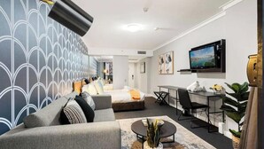 A modern hotel room with a bed, sofa, desk, TV, and a view of the city.