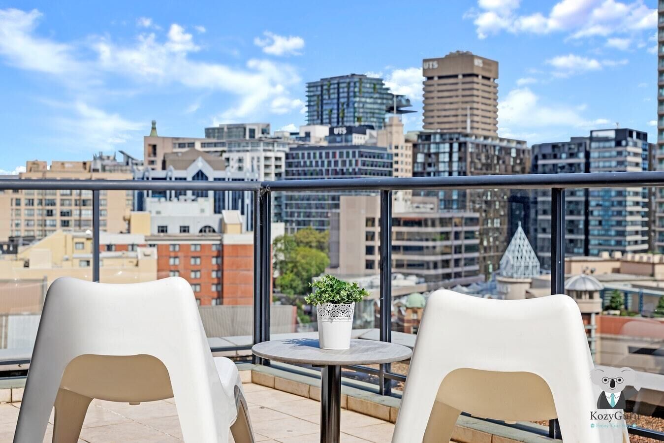 Un balcone con due sedie bianche e un tavolino con una pianta in vaso, con vista su un panorama urbano con diversi edifici.