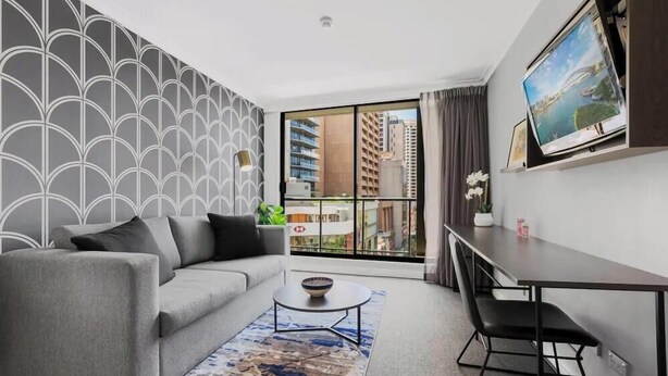 Una sala de estar moderna con un sofá gris, una mesita de centro redonda, un televisor sobre un mueble bajo y un ventanal con vistas a la ciudad.