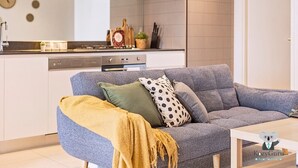 Ein modernes Wohnzimmer mit einem grauen Sofa, einer gelben Decke und einem Kissen mit Punkten. Ein Couchtisch steht davor.