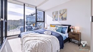 Ein modernes Schlafzimmer mit großem Fenster, einem Bett mit blau-weißer Bettwäsche, einem Nachttisch mit Lampe und zwei Wandbildern.