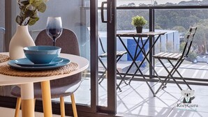 Ein moderner Essbereich mit einem für zwei gedeckten Tisch, einer Vase mit Grünpflanzen und einem Weinglas. Draußen ein Balkon mit Tisch und Stühlen, ein kleines Topfpflanzen und Blick auf die Stadtlandschaft.