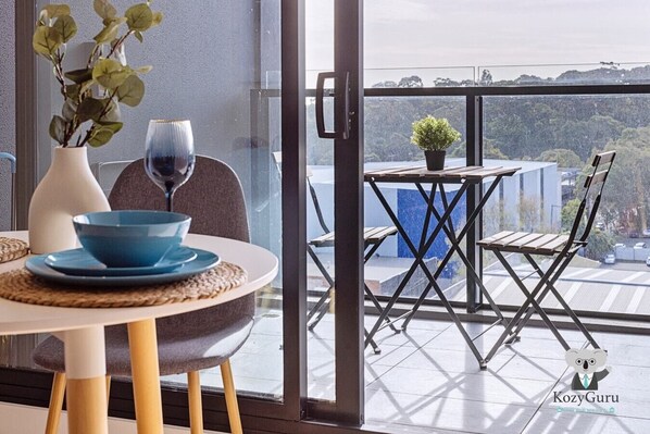 Un espace repas moderne avec une table dressée pour deux, un vase agrémenté de verdure et un verre à vin. À l’extérieur, on trouve un balcon avec une table et des chaises, une petite plante en pot et une vue sur le paysage urbain.