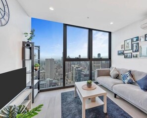 Enjoy a panoramic city view from the comfort of this stylish living room, featuring a sofa, coffee table, flat-screen TV, and a bookshelf with decorative items.
