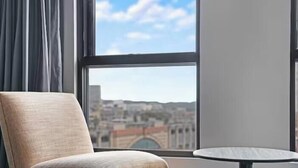 A room with a view of a cityscape through a window, a chair, and a small round table. - KozyGuru | Sydney CBD | High Level 2 Bed Studio (Haymarket)