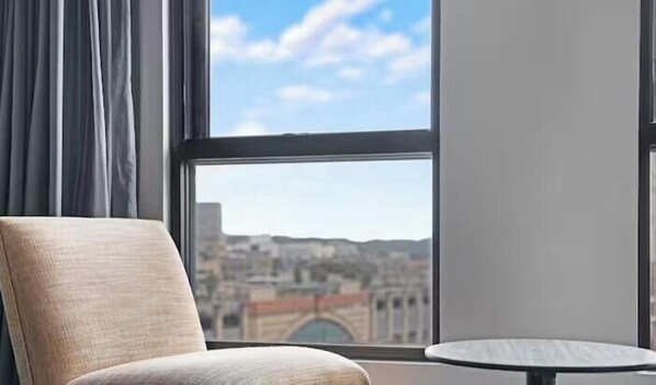 A room with a view of a cityscape through a window, a chair, and a small round table. - KozyGuru | Sydney CBD | High Level 2 Bed Studio (Haymarket)