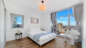 Un dormitorio moderno con una cama grande, una mesita de noche, una silla, una ventana con cortinas y vistas a la ciudad.