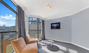 Et moderne værelse med sofa, lille bord, fjernsyn og store vinduer med gardiner.