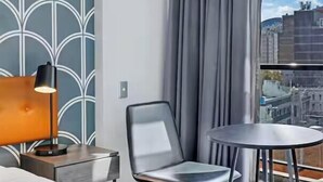 Une chambre d’hôtel au design moderne, dotée d’une grande fenêtre avec des rideaux, d’une table ronde, d’une chaise et d’une lampe sur une table d’appoint.