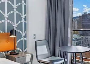 Et hotelværelse med moderne design, med et stort vindue med gardiner, et rundt bord, en stol og en lampe på et sengebord.