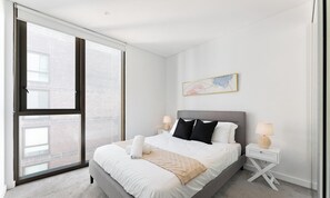 A modern hotel room with a large bed, two bedside tables, a window with blinds, and a wall-mounted artwork.
