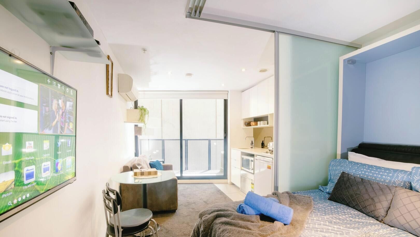 Quarto de hotel moderno com varanda, escrivaninha, cama com roupa de cama estampada e uma tela grande exibindo uma interface de mídia social.
