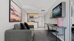 A modern living room with a grey sofa, a flat-screen TV mounted on the wall, a kitchen area in the background, and a small dining table with chairs. - KozyGuru | Haymarket | Bright City Studio (Haymarket)