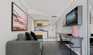 Un soggiorno moderno con un divano grigio, una TV a schermo piatto montata a parete, un angolo cottura sullo sfondo e un piccolo tavolo da pranzo con sedie.