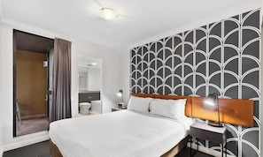 A hotel room with a bed, a nightstand, a lamp, a bathroom with a toilet and mirror, and a door leading to an outdoor area.