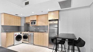 A modern kitchen with wooden cabinets, a stainless steel refrigerator, a microwave, a washing machine, and a dining area with a table and chairs. - KozyGuru | Haymarket | High Level 2B APT (Haymarket)