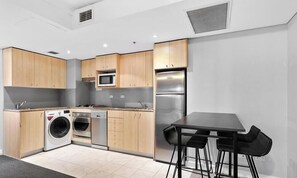 A modern kitchen with wooden cabinets, a stainless steel refrigerator, a microwave, a washing machine, and a dining area with a table and chairs.