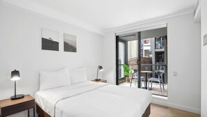 Un dormitorio con cama, lámparas de noche y vistas a un balcón exterior con sillas y una planta.