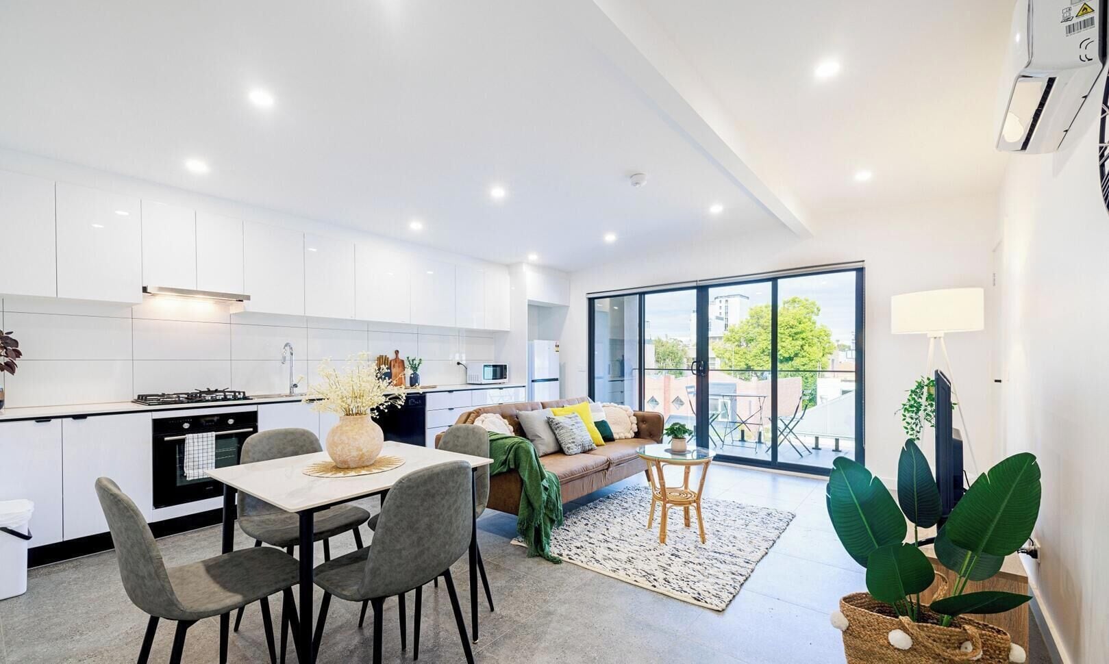 Une cuisine moderne avec des meubles blancs, un coin repas comprenant une table et des chaises, un canapé et une grande porte-fenêtre coulissante donnant sur un balcon.