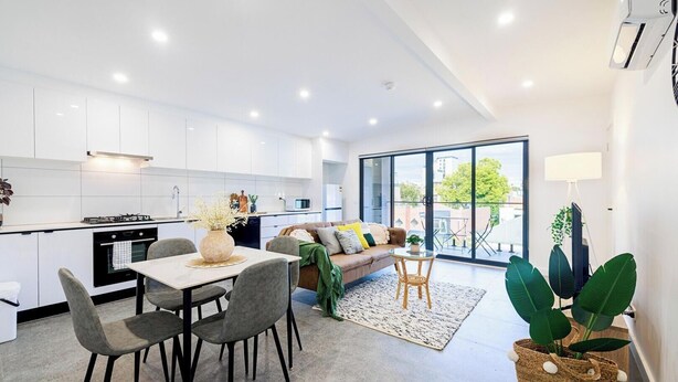 A modern kitchen with white cabinets, a dining area with a table and chairs, a sofa, and a large sliding glass door leading to a balcony. - KozyGuru | Richmond | Cozy Unit and Parking near CBD (Cremorne)