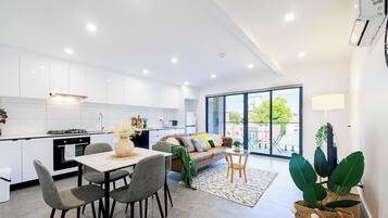 Une cuisine moderne avec des meubles blancs, un coin repas comprenant une table et des chaises, un canapé et une grande porte-fenêtre coulissante donnant sur un balcon.