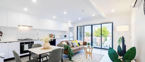 Une cuisine moderne avec des meubles blancs, un coin repas comprenant une table et des chaises, un canapé et une grande porte-fenêtre coulissante donnant sur un balcon.