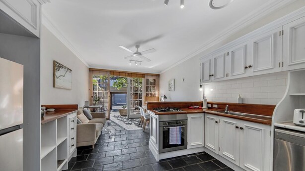 A modern kitchen with white cabinetry, a central island, and a dining area with a sofa. - KozyGuru | Glebe | Cozy 2 Bedroom Terrace House (Glebe)
