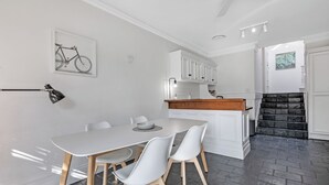 A modern kitchen with white cabinetry, a dining area with a white table and chairs, and a staircase with dark tiles. - KozyGuru | Glebe | Cozy 2 Bedroom Terrace House (Glebe)