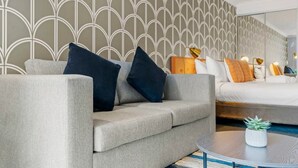 A modern hotel room with a grey sofa, a bed with multiple pillows, a small round table with a plant, and a patterned wallpaper.