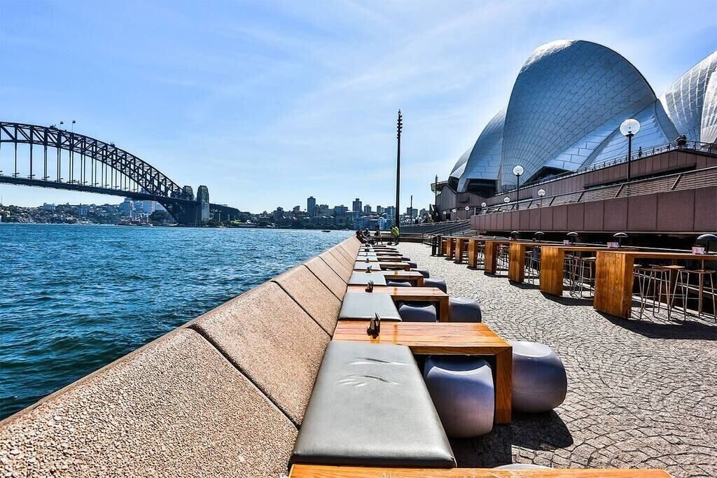 Eine Uferpromenade mit Sitzgelegenheiten und Blick auf das Sydney Opera House und die Harbour Bridge.