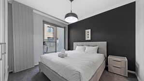 A bedroom with a bed, a bedside table, a hanging light, and a view through a window. - KozyGuru | Ryde | Cosy 2Bed Apt with Private Yard (Ryde)
