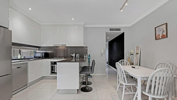 A modern kitchen with white cabinetry, a central island, and a dining area with white chairs and a table. - KozyGuru | Ryde | Cosy 2Bed Apt with Private Yard (Ryde)