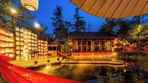 Un bâtiment traditionnel en bois, avec un étang devant, entouré d’arbres et illuminé par des lumières chaleureuses.